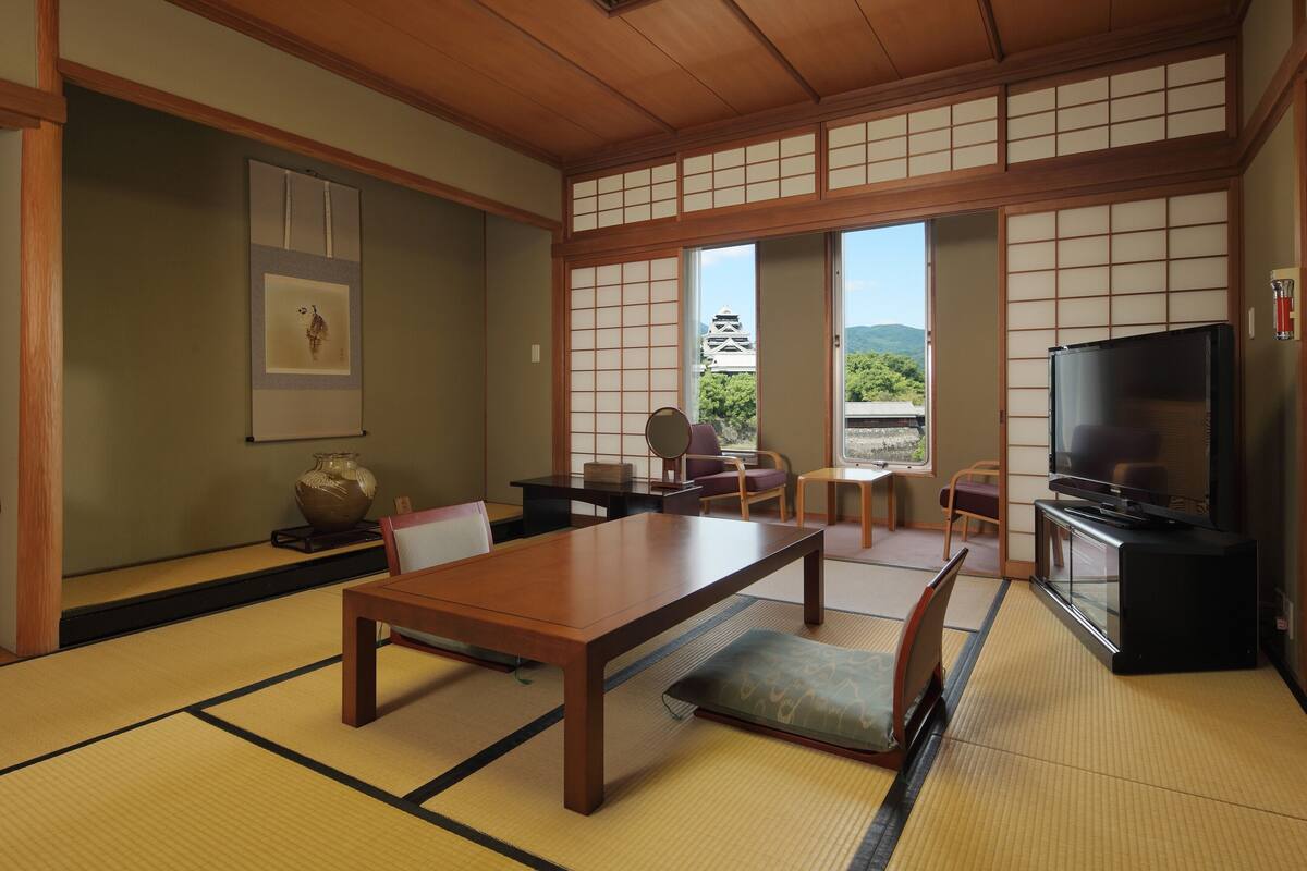 A traditional Japanese room with tatami flooring, a low table, chairs, a television, and a view of a pagoda through large windows.