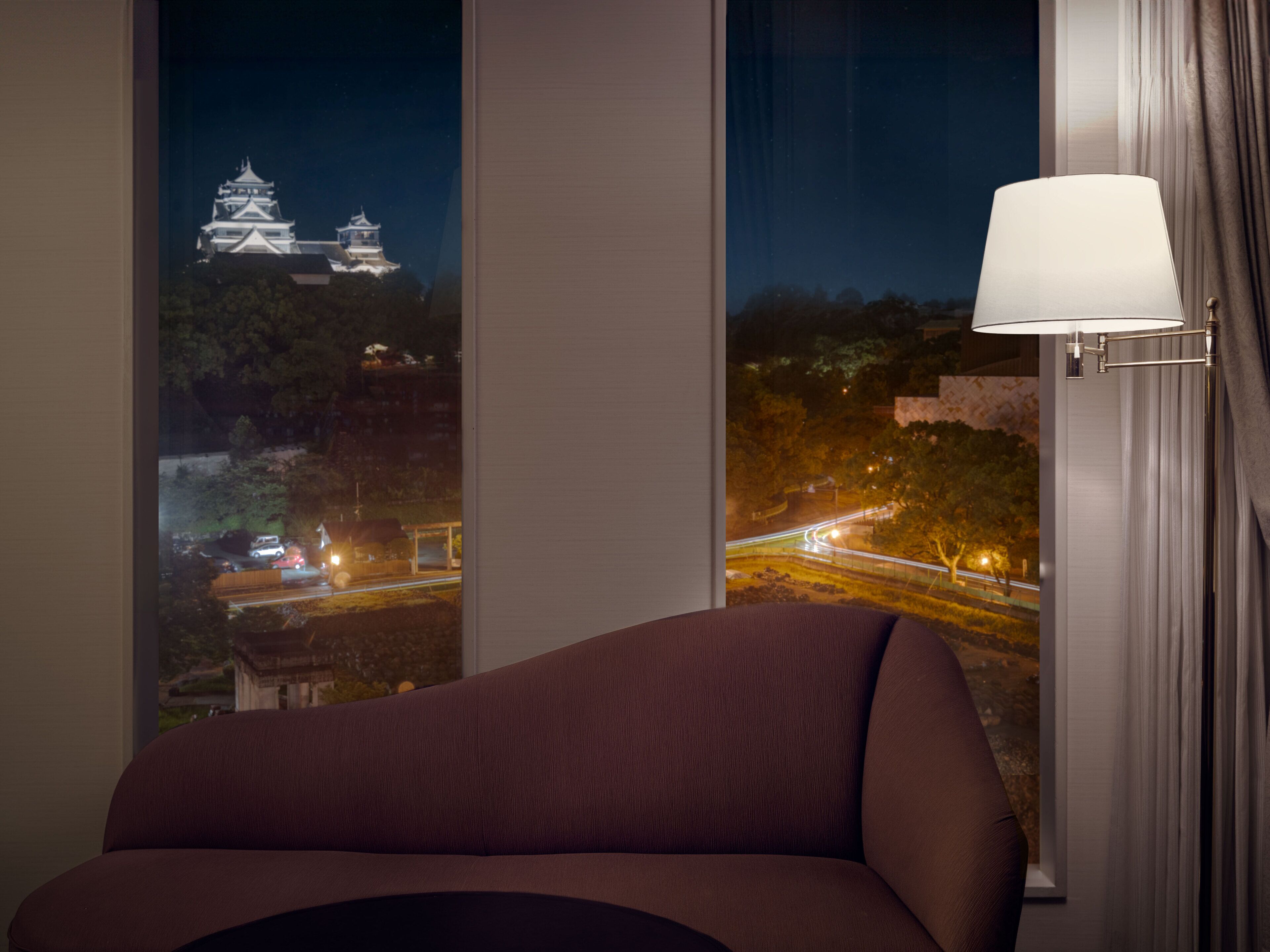 A room with a view of a traditional Japanese castle at night, a modern lamp, and a sofa.