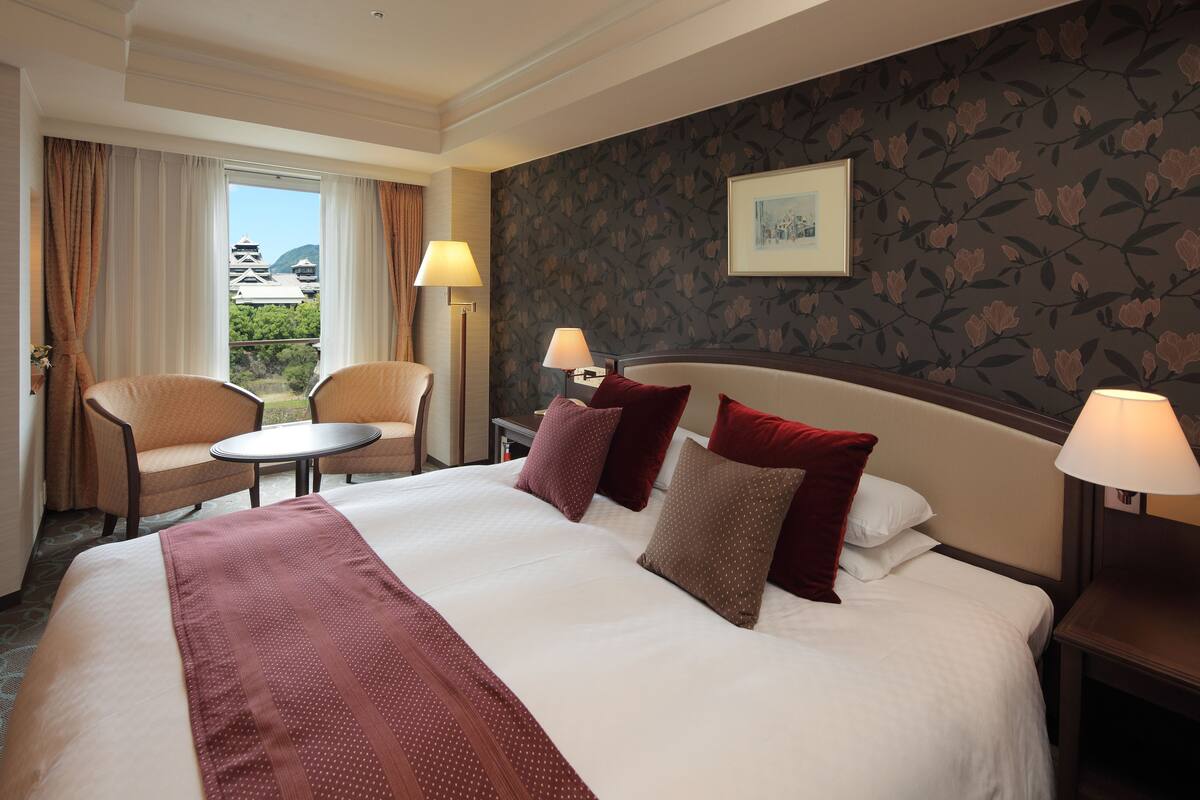 A hotel room with a large bed, two chairs, a small table, and a view of a traditional building.