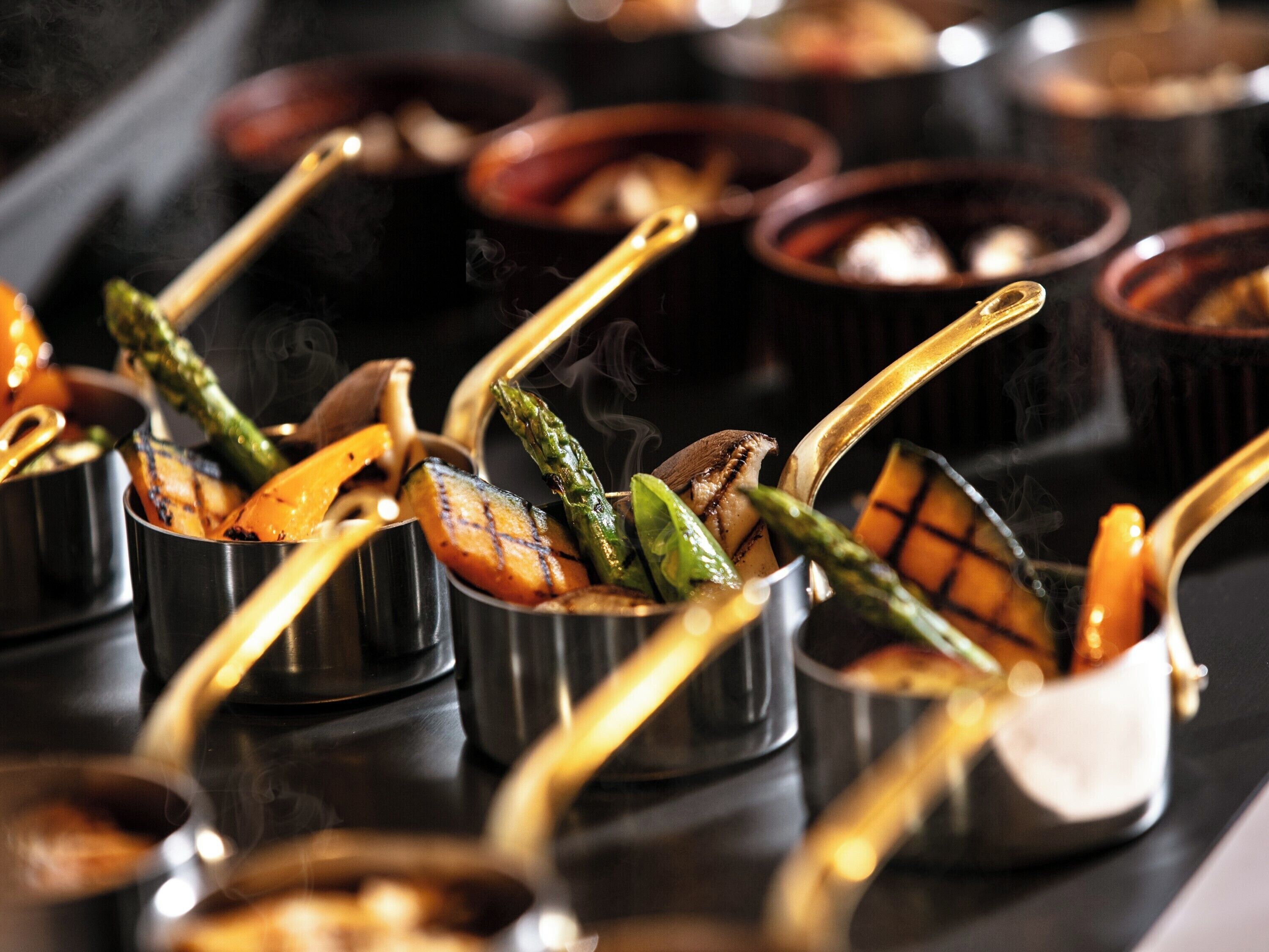 A buffet with various dishes, including grilled vegetables and meats, served in metal containers with serving utensils.