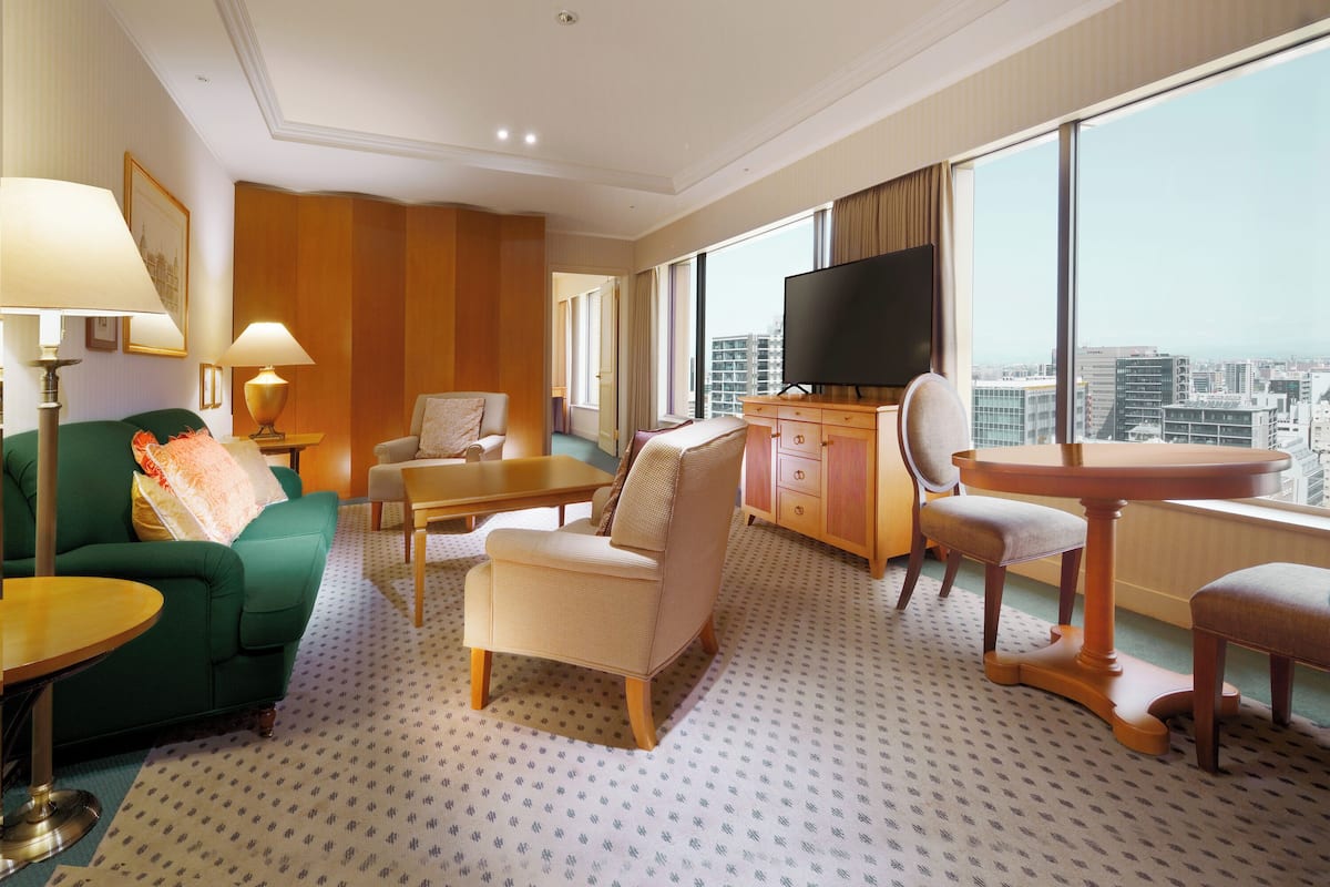 Une chambre d’hôtel avec un canapé vert, un meuble en bois doté d’un téléviseur à écran plat, une petite table et une vue sur la ville à travers de grandes fenêtres.