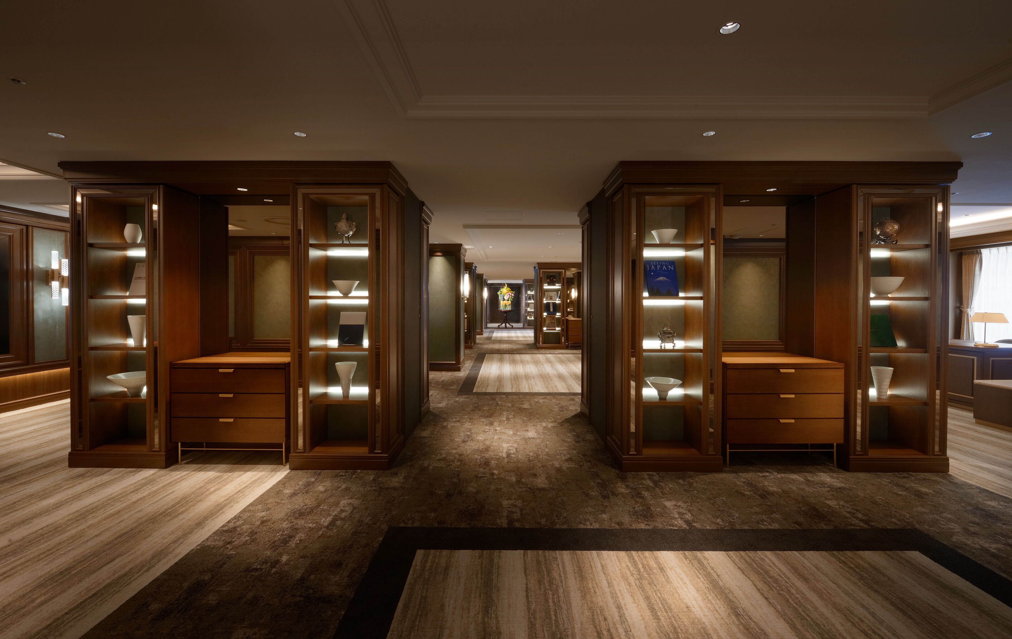 A spacious hotel lobby with a central carpeted area, flanked by wooden bookcases with glass doors.
