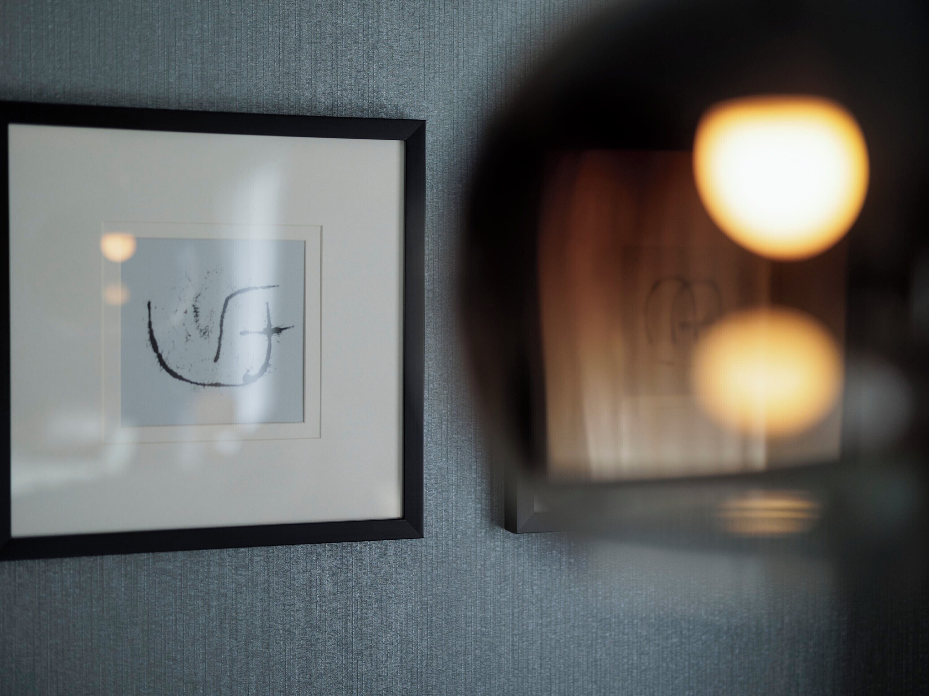 A framed artwork with a simple line drawing on a textured grey wall.