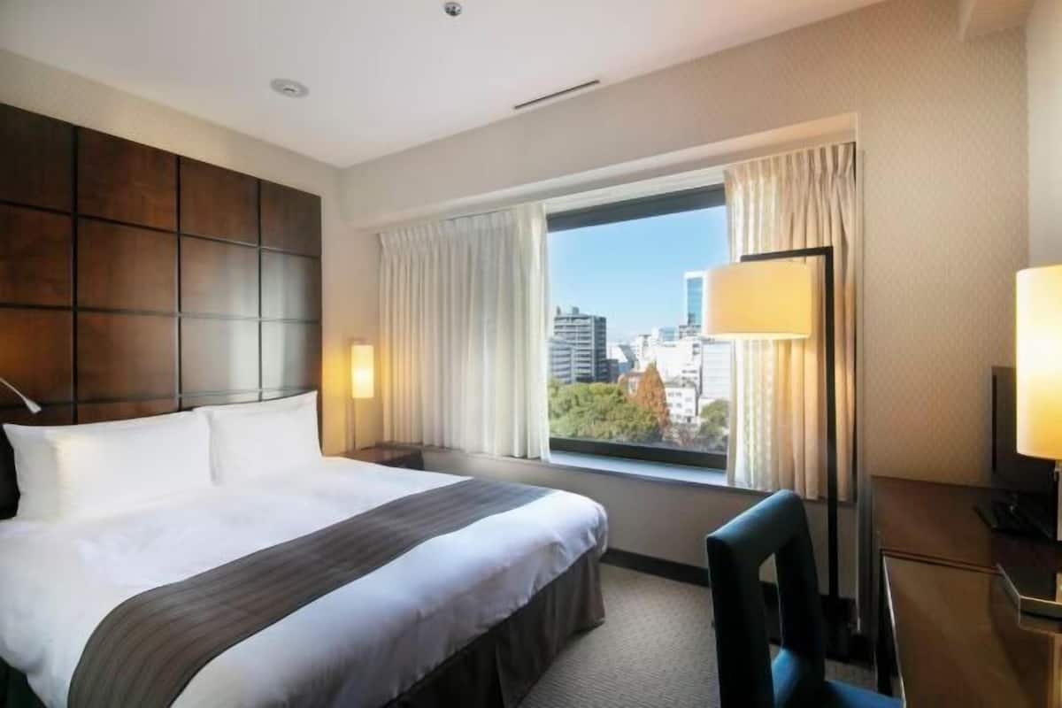 Une chambre d’hôtel avec un grand lit, un bureau, une chaise et une fenêtre donnant sur des immeubles.