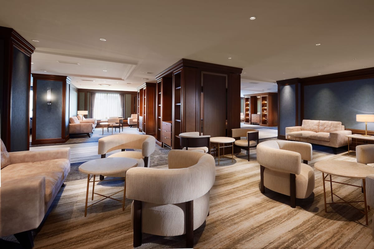 Un hall d’hôtel spacieux, doté de plusieurs espaces de détente, notamment des canapés et des fauteuils, d’une grande armoire en bois et d’un plafond bien éclairé grâce à des spots encastrés.