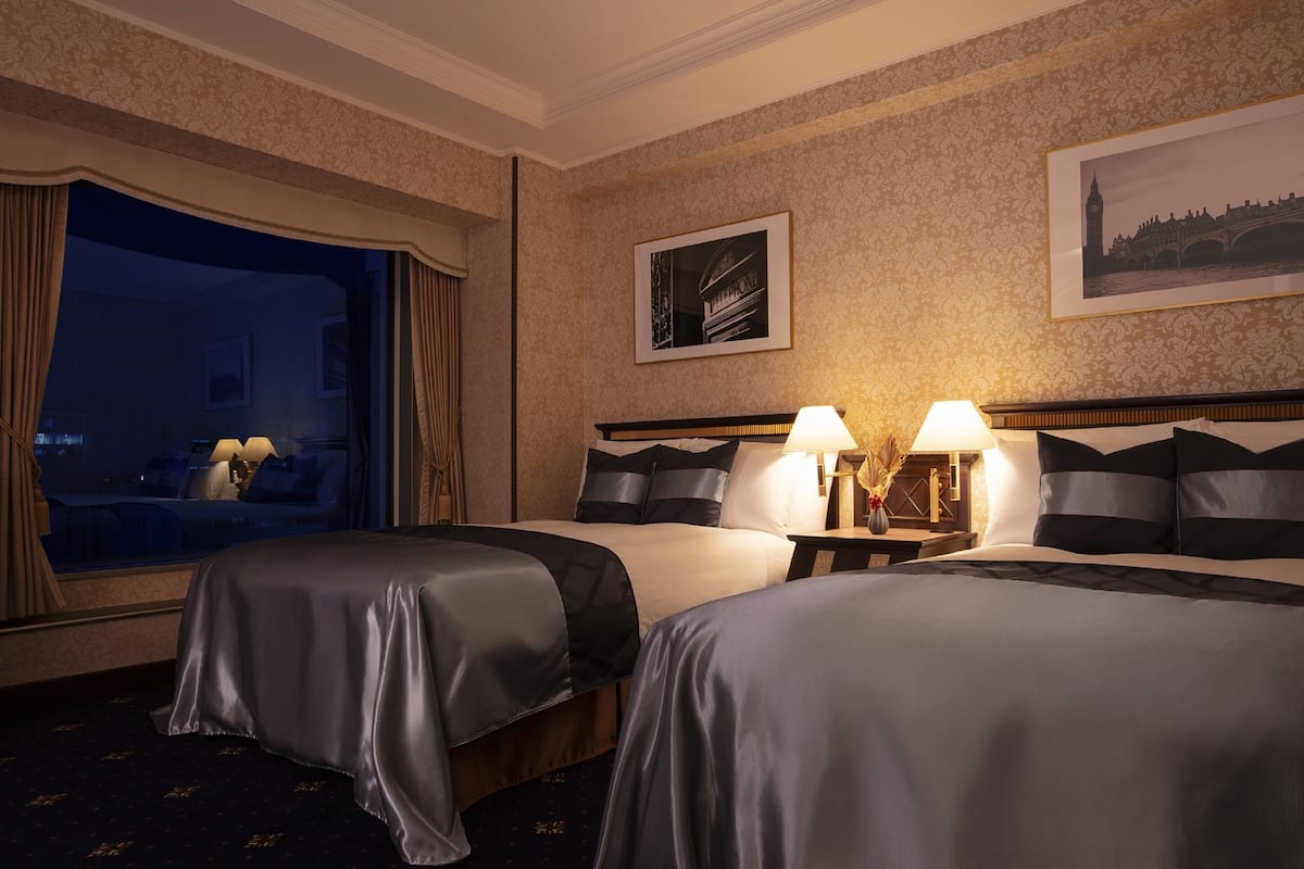 Une chambre d’hôtel avec deux lits, une grande fenêtre avec des rideaux, un tableau encadré au mur et une vue nocturne sur l’extérieur.