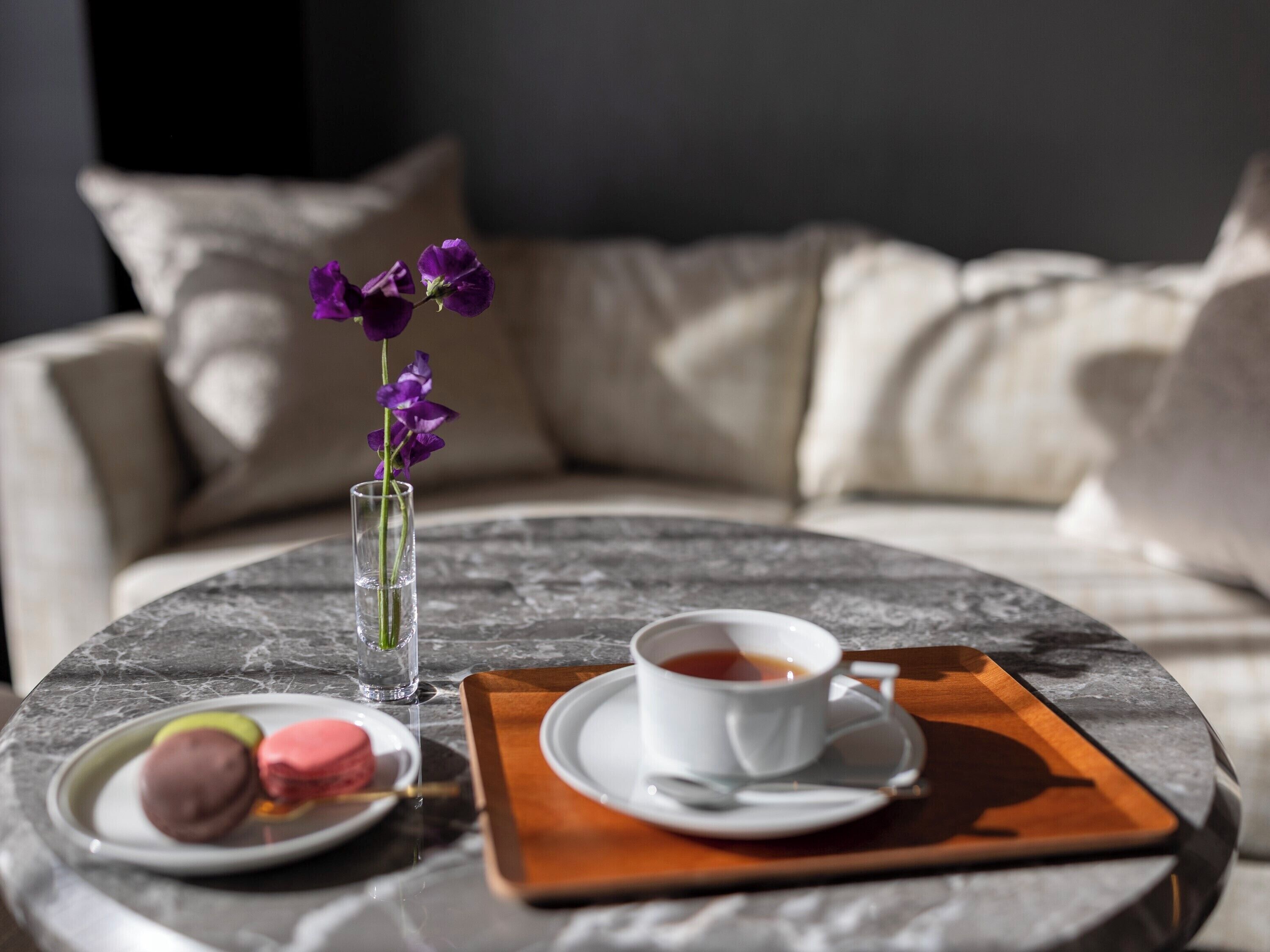 A round marble table with a cup of tea, a small vase with purple flowers, and a plate with macarons.