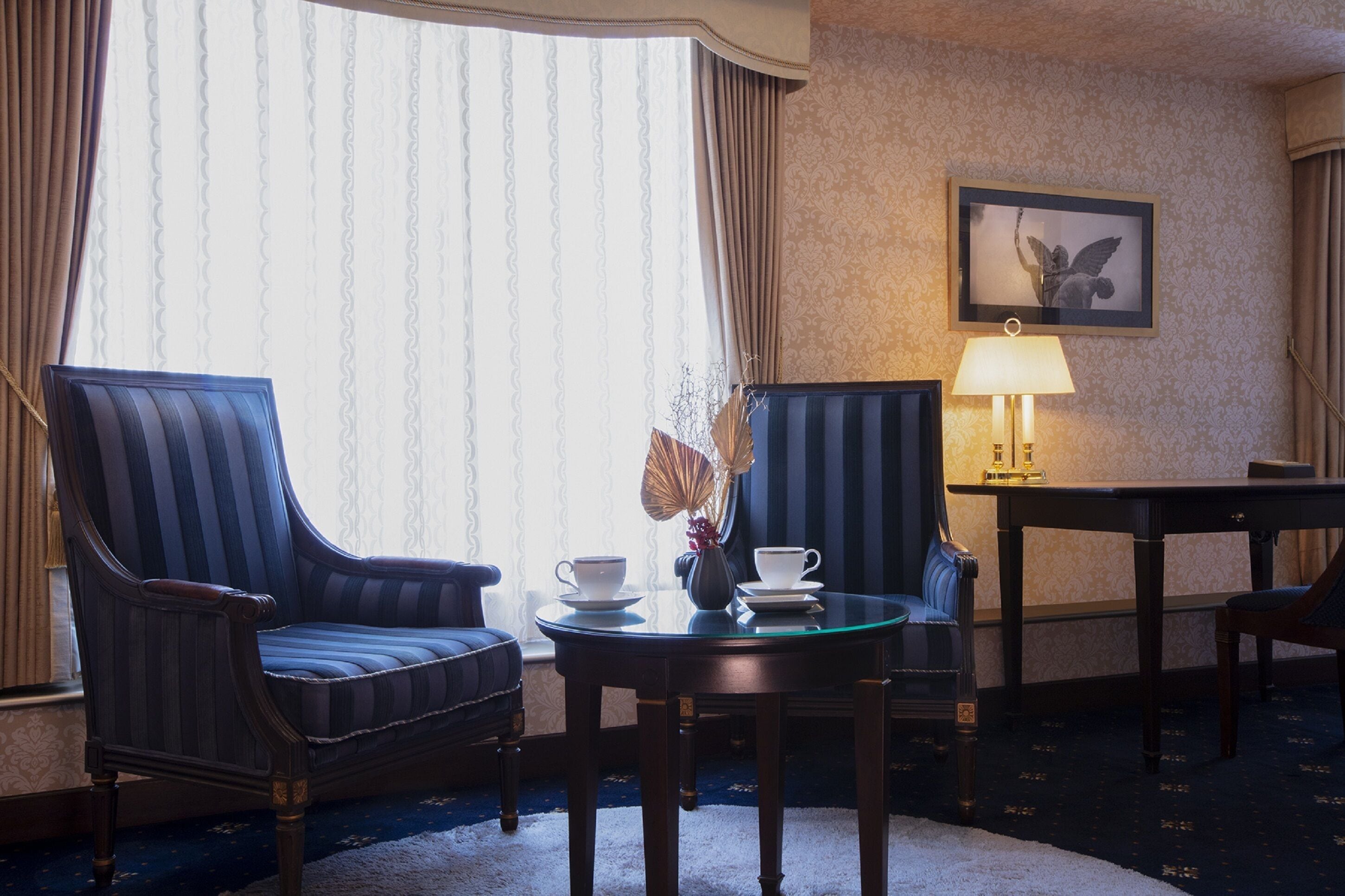 A hotel room with two upholstered armchairs, a small round table with a vase and a cup, and a lamp on a side table.