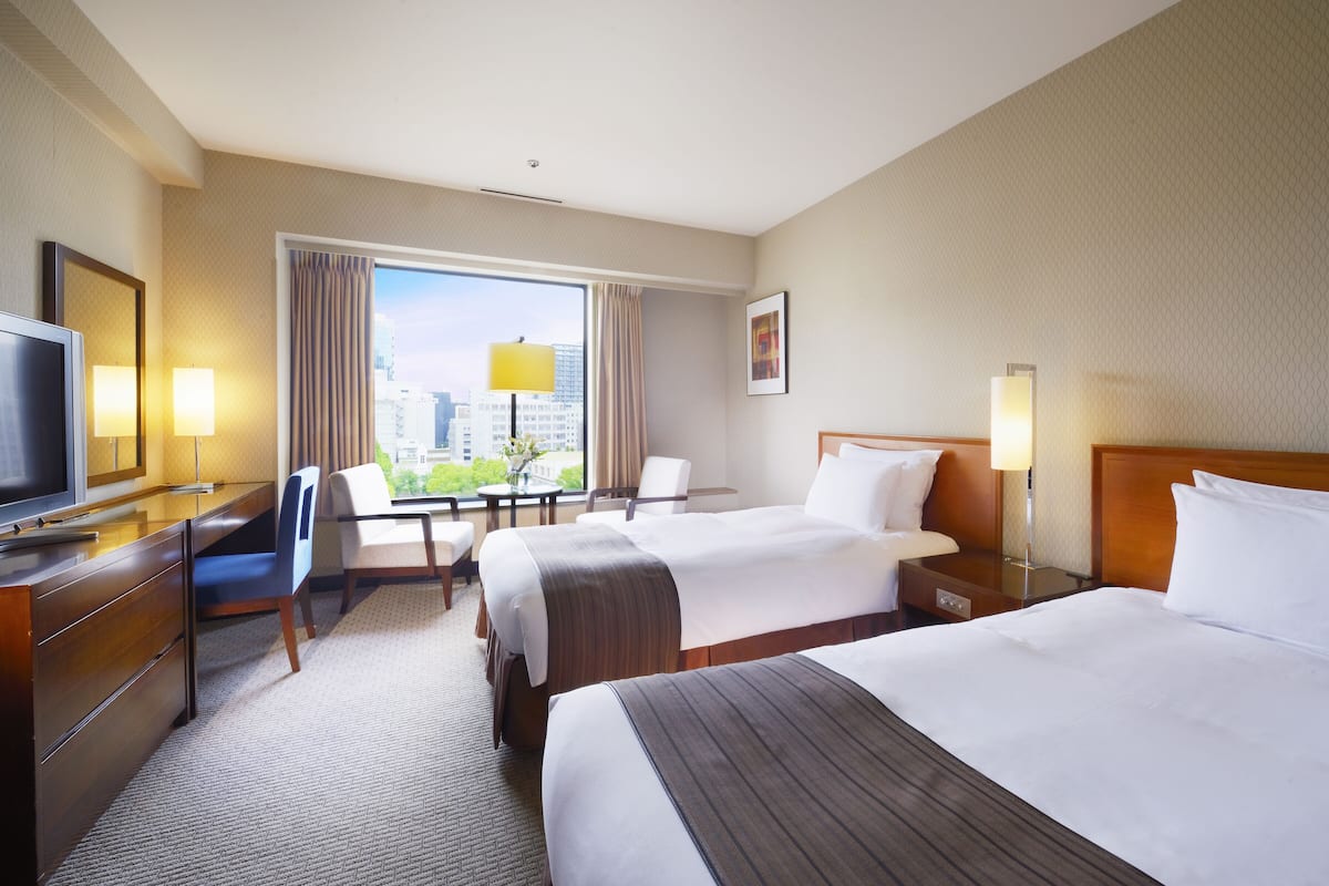 Une chambre d’hôtel avec deux lits, un bureau avec une télévision, une chaise, une table avec une lampe et une fenêtre donnant sur la ville.