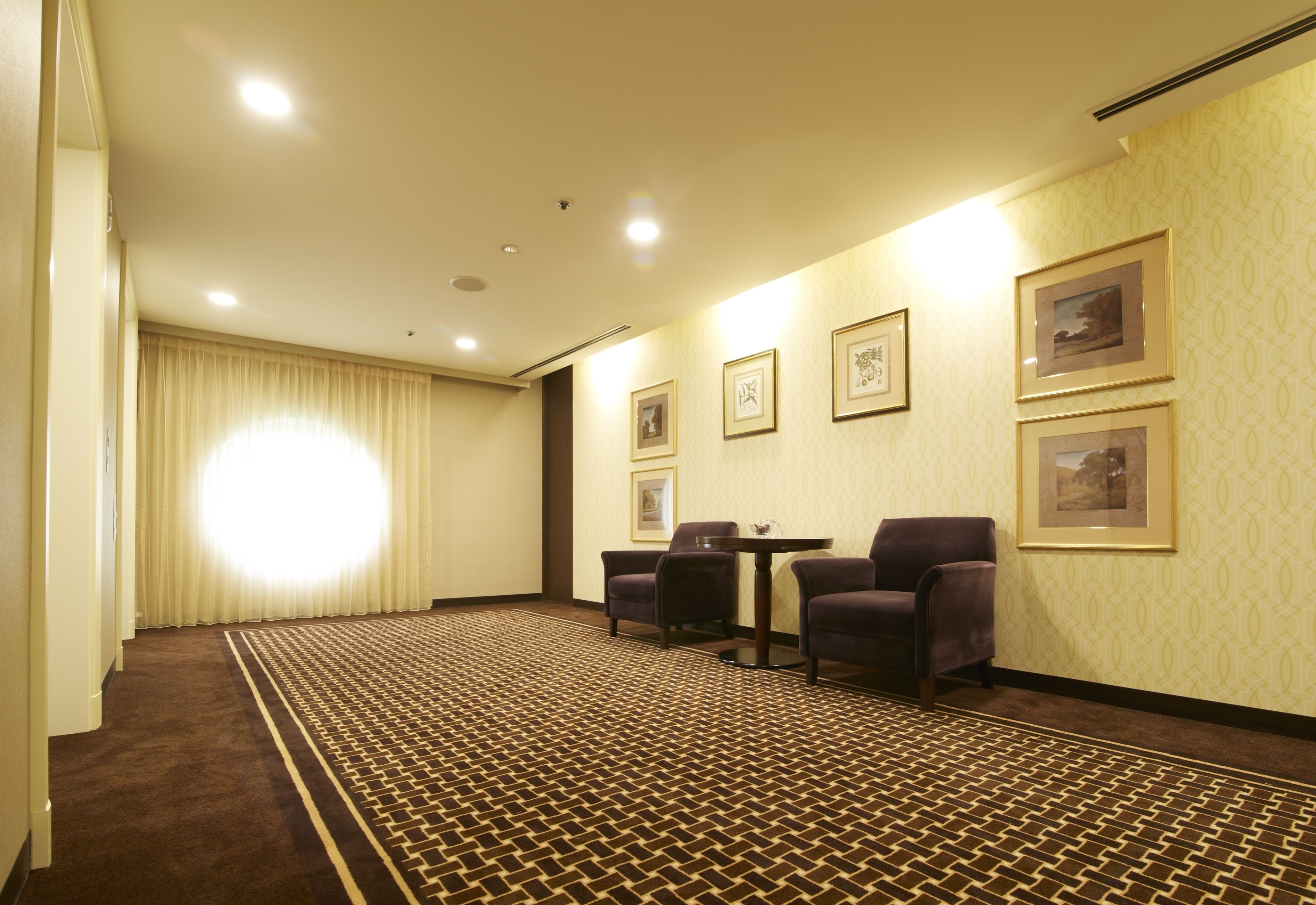 A hotel room with a patterned carpet, two chairs, a small table, and framed pictures on the wall.