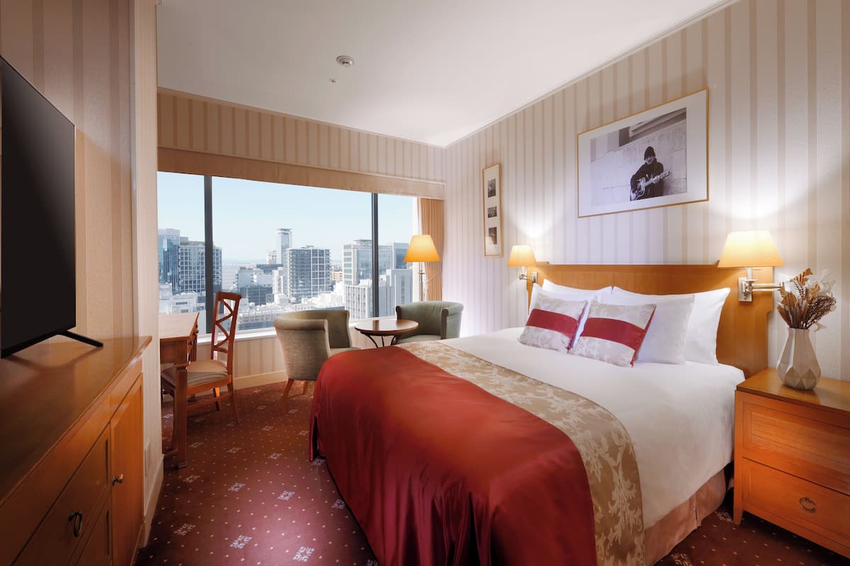 Une chambre d’hôtel avec un grand lit, deux fauteuils, une petite table, un téléviseur et une vue sur la ville.