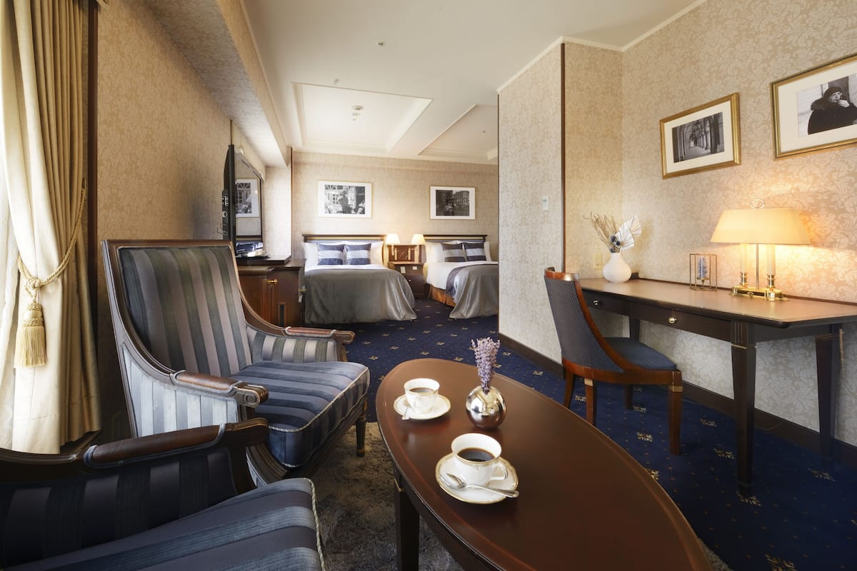 Une chambre d’hôtel comprenant un lit, deux fauteuils, un bureau avec une lampe et une petite table avec des tasses et un vase.