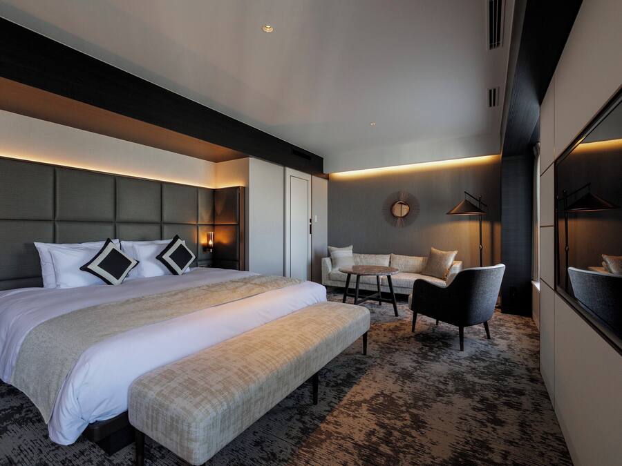 A modern hotel room with a large bed, a sofa, a small table, and a bench.