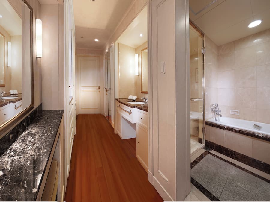 A hotel hallway with a wooden floor, a bathroom with a bathtub, and a mirror reflecting the bathroom.