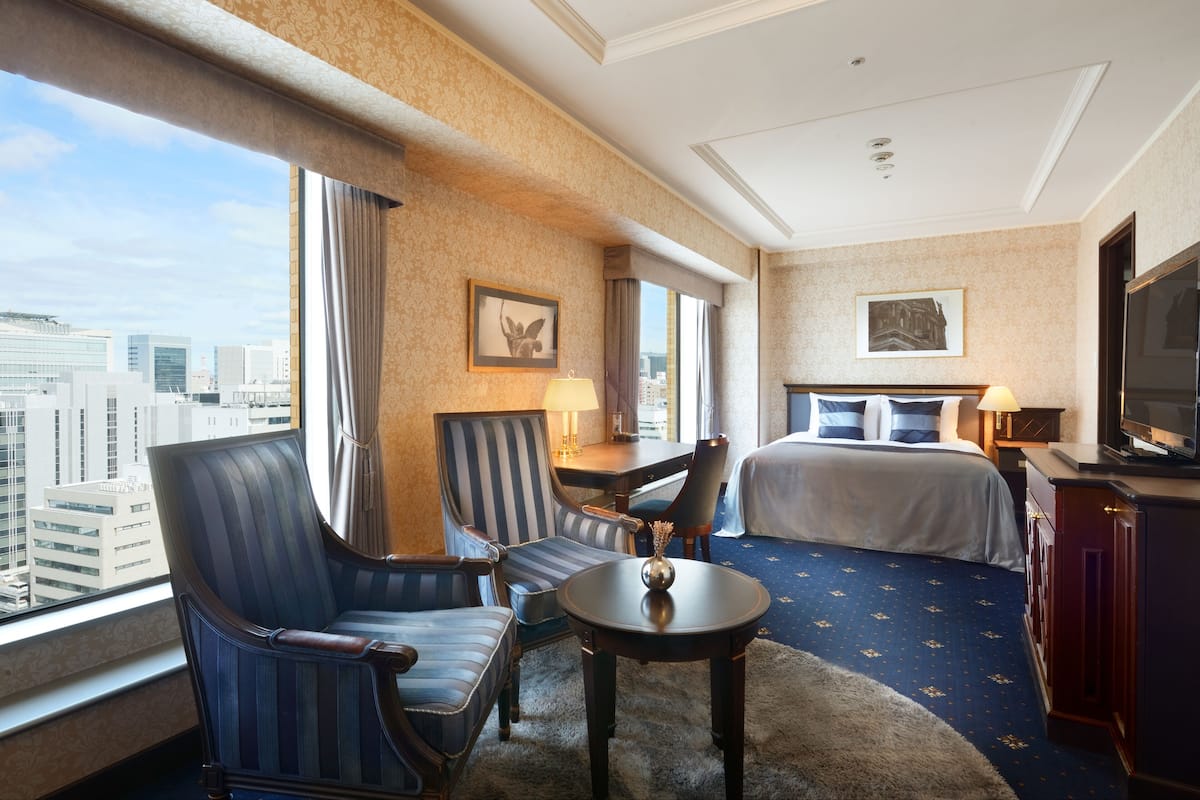 Une chambre d’hôtel avec une grande fenêtre, un lit, deux fauteuils, une petite table et un téléviseur.