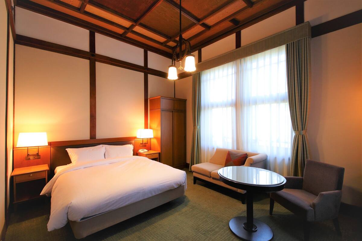 A hotel room with a bed, two bedside lamps, a sofa, a round table, and a window with curtains.