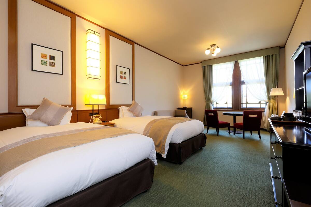 A hotel room with two beds, a desk, a chair, a TV, and a window with curtains.