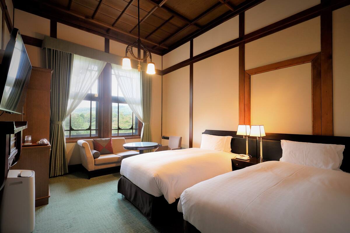 A hotel room with two beds, a TV, a desk, and a view of the outdoors.