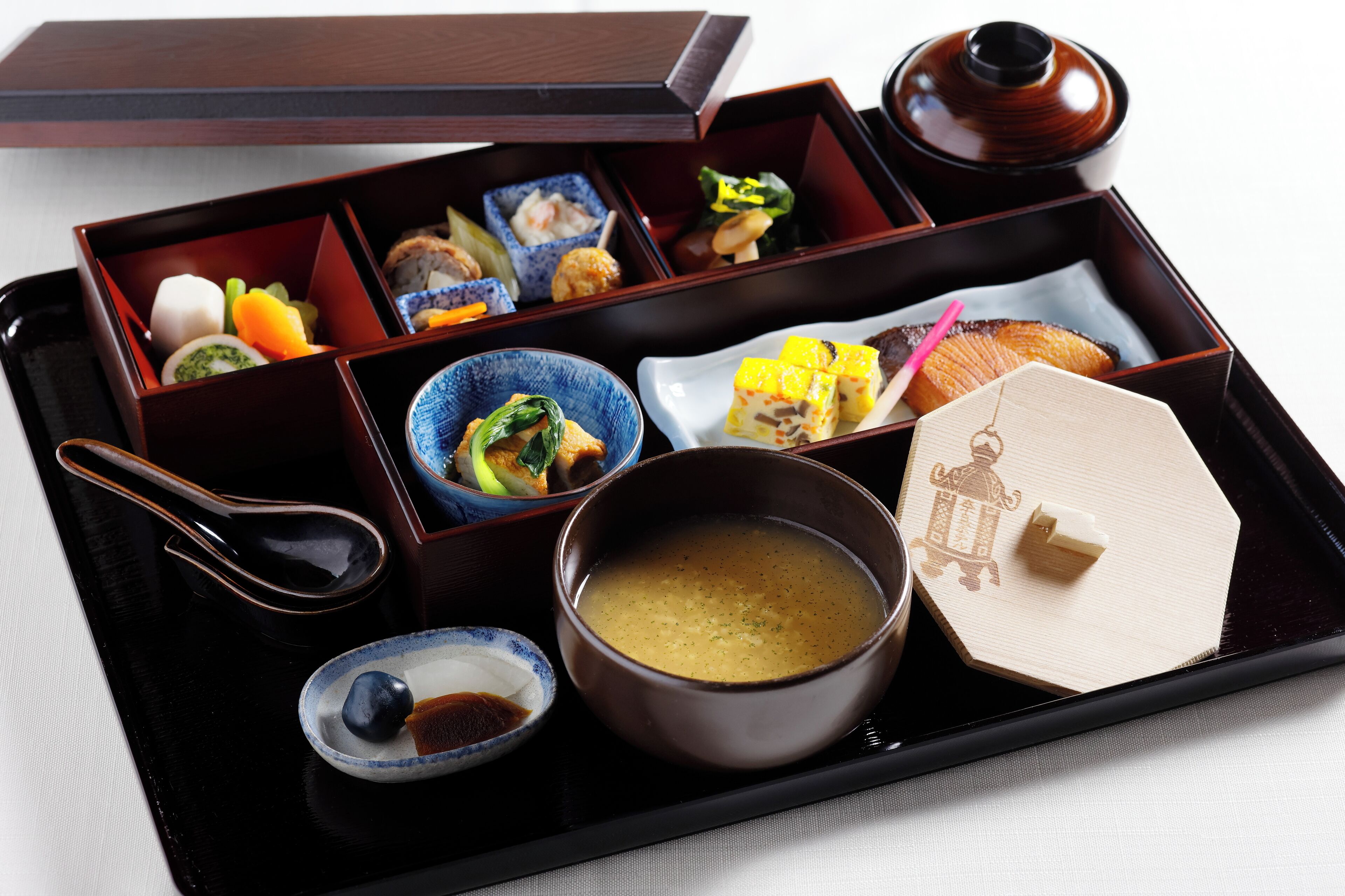 Una caja bento japonesa tradicional con diferentes alimentos y un tazón pequeño de sopa.