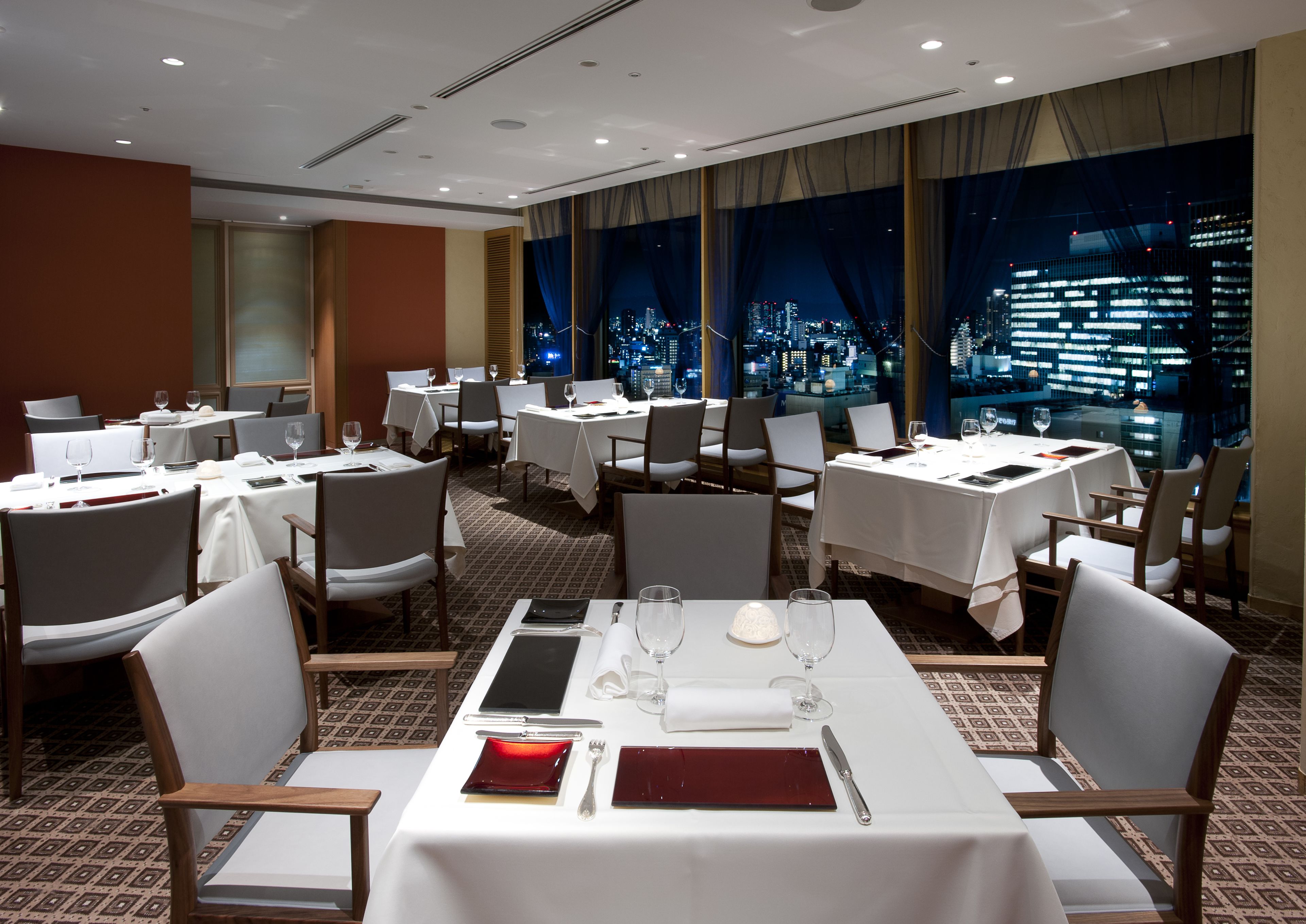 A hotel dining area with multiple tables set for dining, chairs, and a city view through large windows.