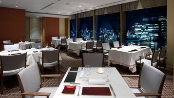 A hotel dining area with multiple tables set for dining, chairs, and a city view through large windows.