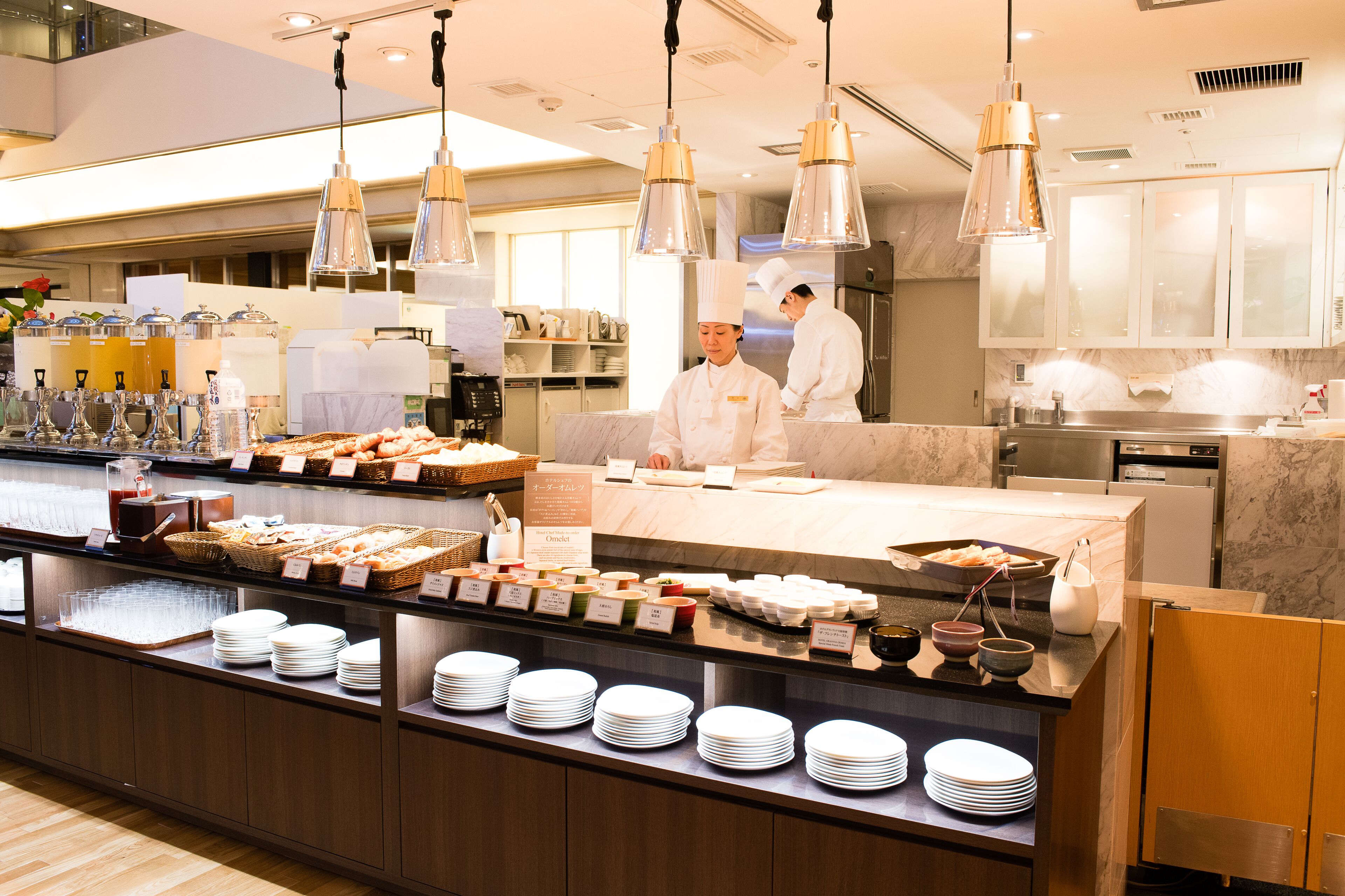 A buffet area with various food items and dishes, a kitchen in the background, and two chefs at work.