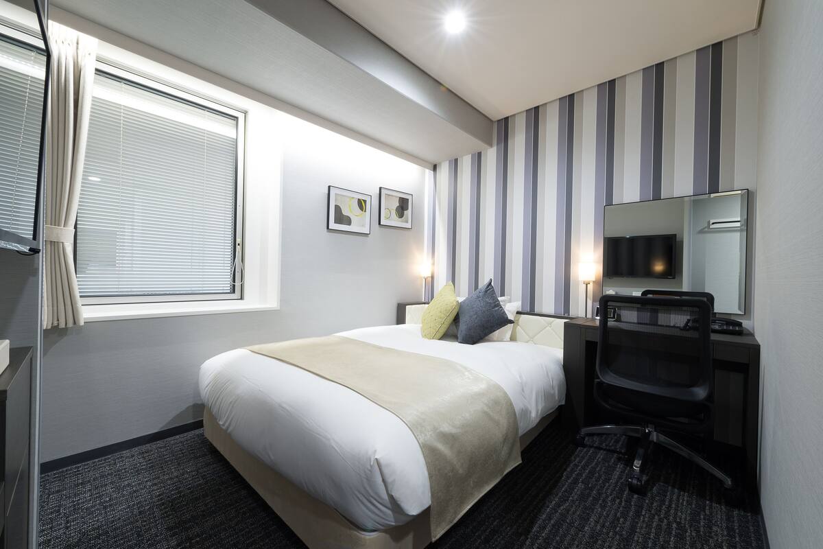 A hotel room with a large bed, a desk with a chair, a window with curtains, and two framed pictures on the wall.
