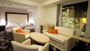 Quarto de hotel com cama, duas poltronas, um sofá, uma mesa pequena e janela com vista para a cidade.