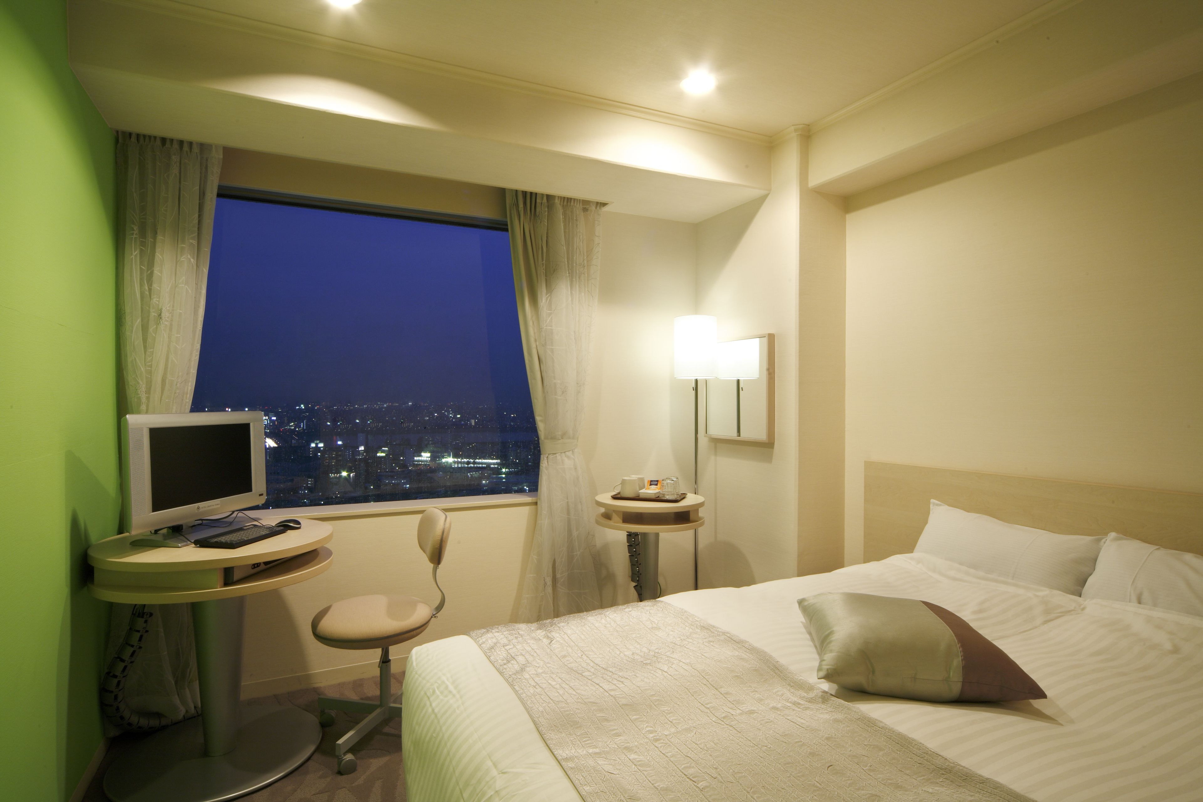 Une chambre d’hôtel avec un lit, un bureau avec un ordinateur, une chaise, une table de chevet avec une lampe et une fenêtre donnant sur la ville la nuit.