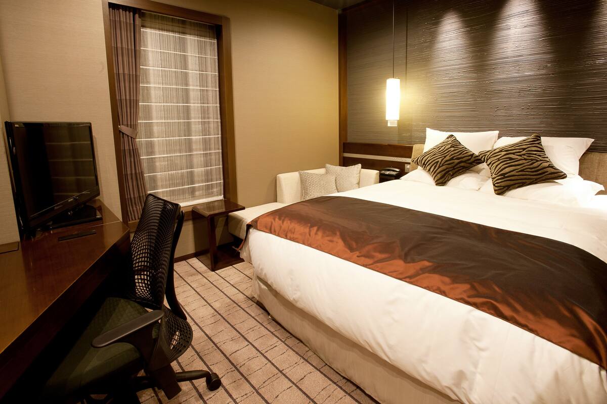 A hotel room with a large bed, a desk with a chair, a television, and a window with curtains.
