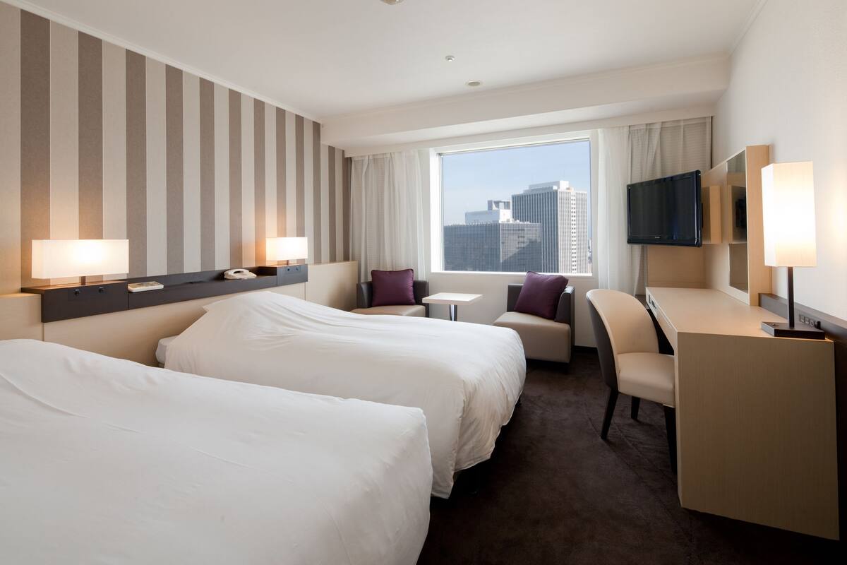 A hotel room with two beds, a desk, a chair, a TV, and a window with a city view.