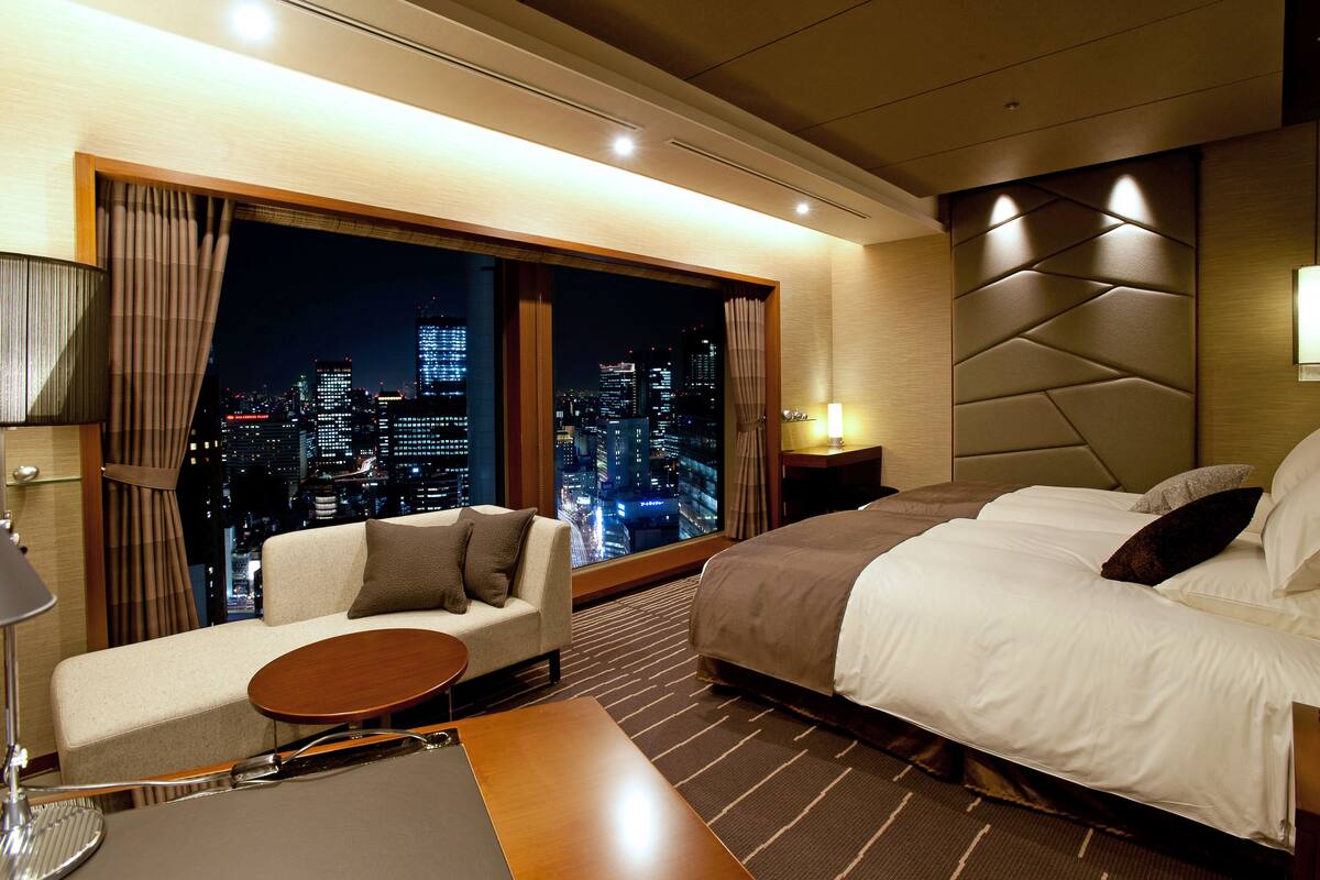 A hotel room with a large window offering a city view, a sofa, a bed, and a small table.