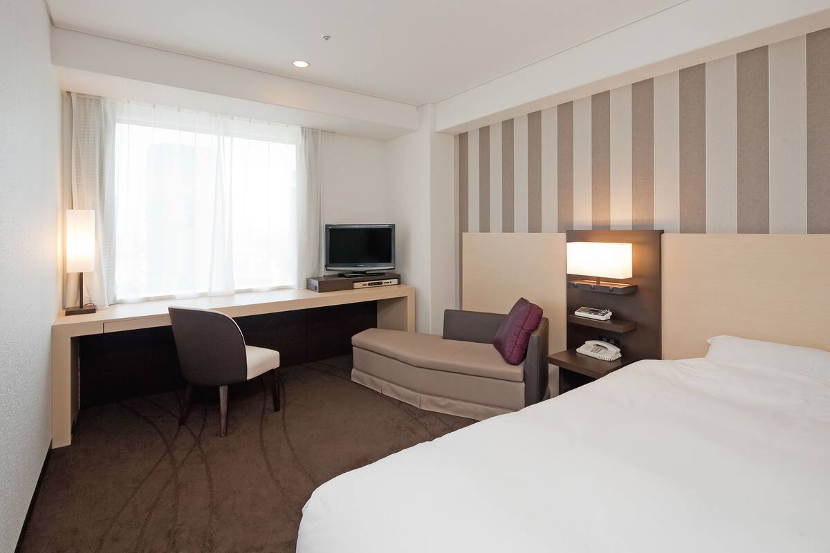 A hotel room with a bed, a desk with a computer, a chair, a TV, and a nightstand with a lamp and phone.