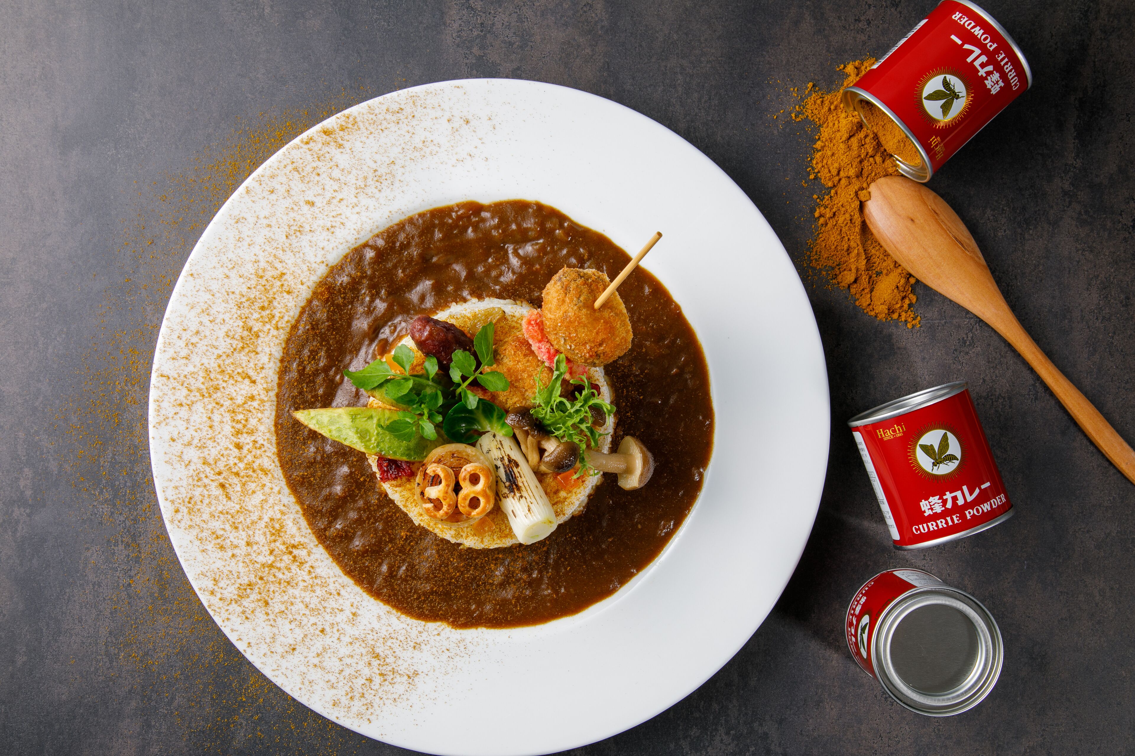 A plate with a dish of curry powder, a wooden spoon, and a can of curry powder.