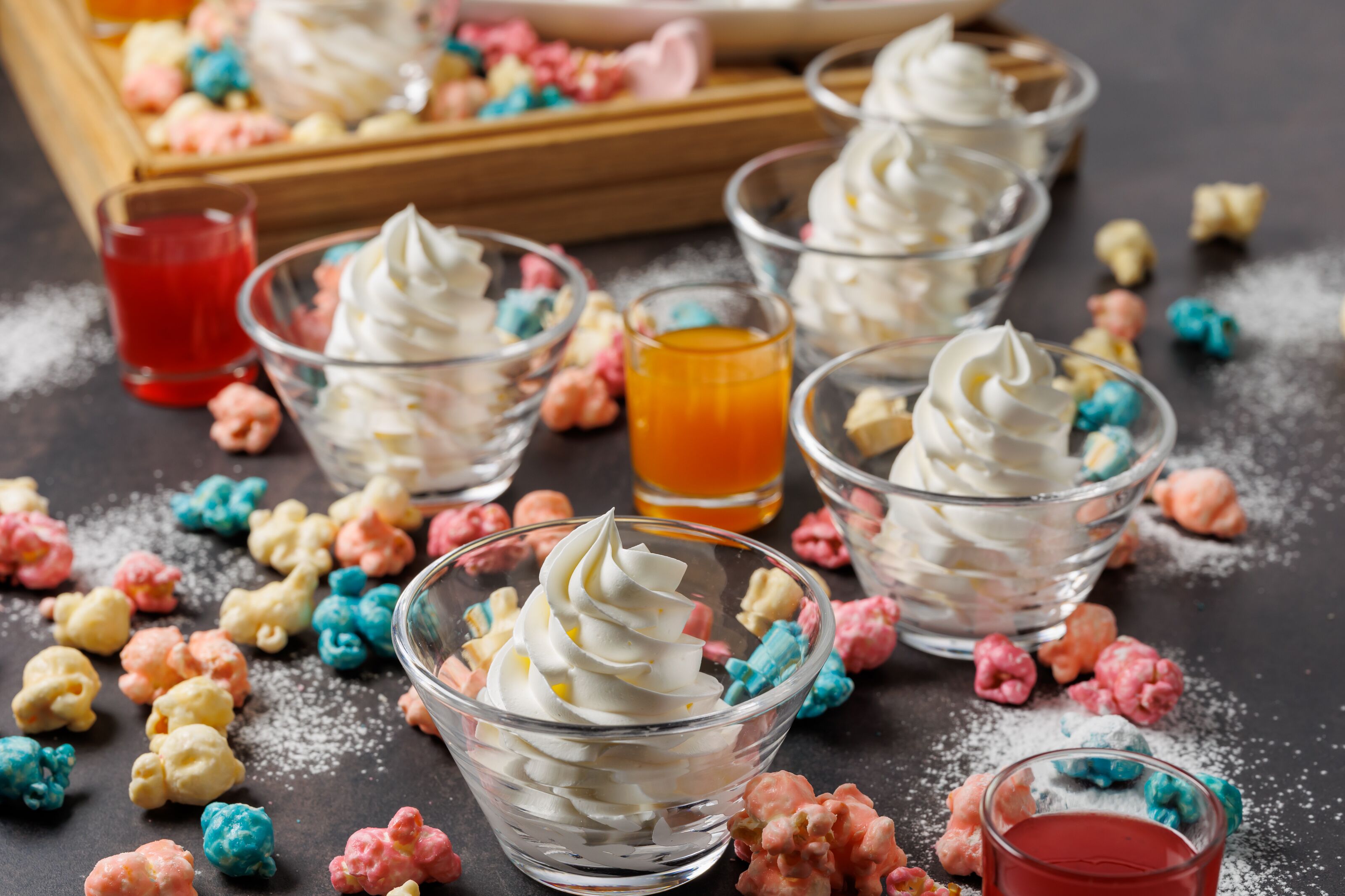 Une table de desserts avec de la crème fouettée, du pop-corn de différentes couleurs et deux verres de jus.