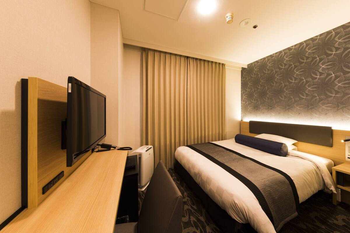 A hotel room with a bed, a desk with a flat-screen TV, a small table, and a wall-mounted lamp.
