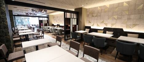A modern restaurant interior with multiple dining tables and chairs, a patterned wall, and a view of the dining area through a glass partition.