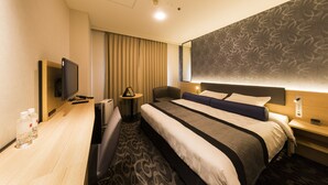 Een hotelkamer met een groot bed, een bureau met een flatscreen televisie, een stoel, een nachtkastje met een lamp en een behangen muur.