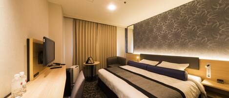 A hotel room with a large bed, a desk with a flat-screen TV, a chair, a bedside table with a lamp, and a patterned wall.