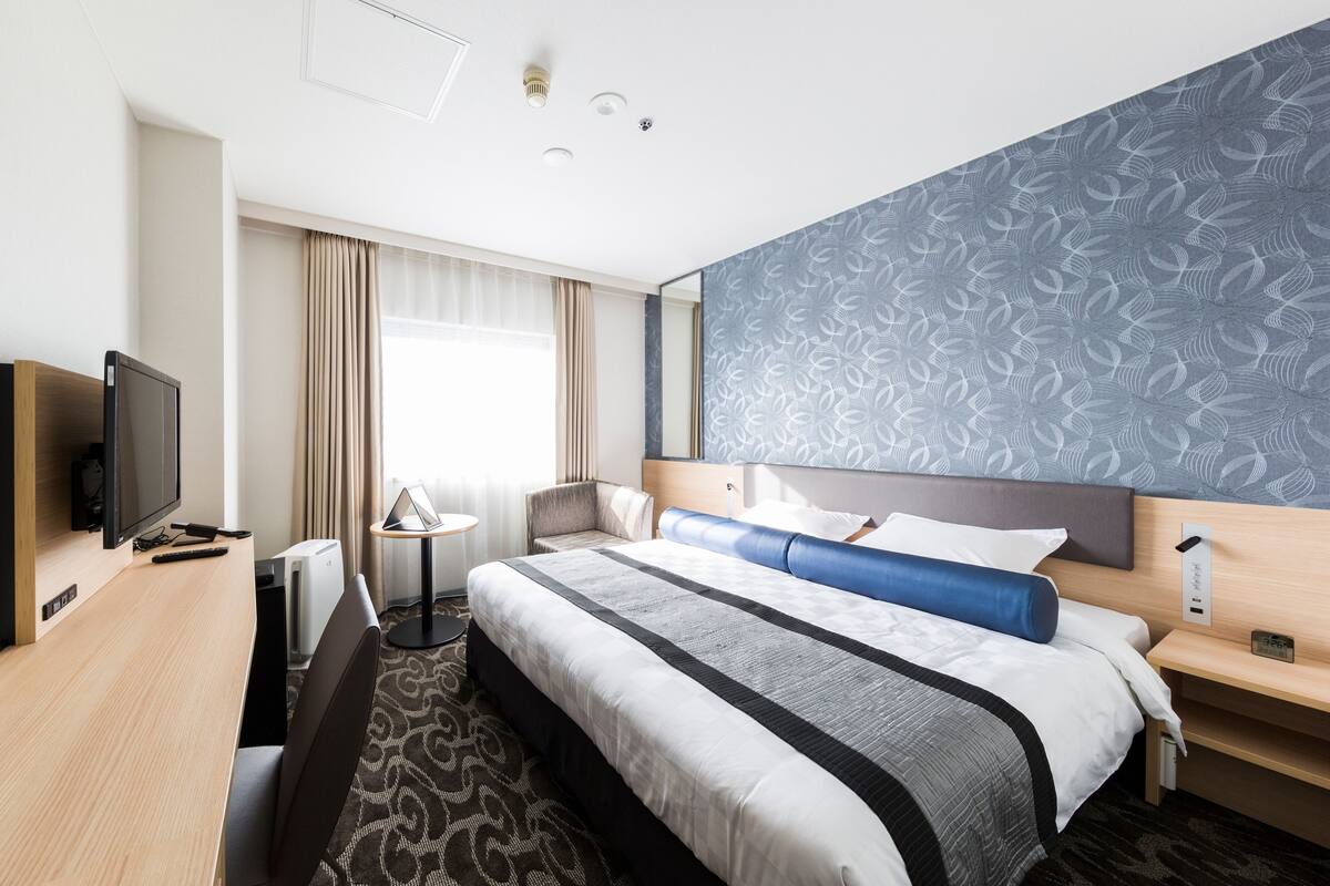 A hotel room with a large bed, a desk with a flat-screen TV, a bedside table, and a window with curtains.