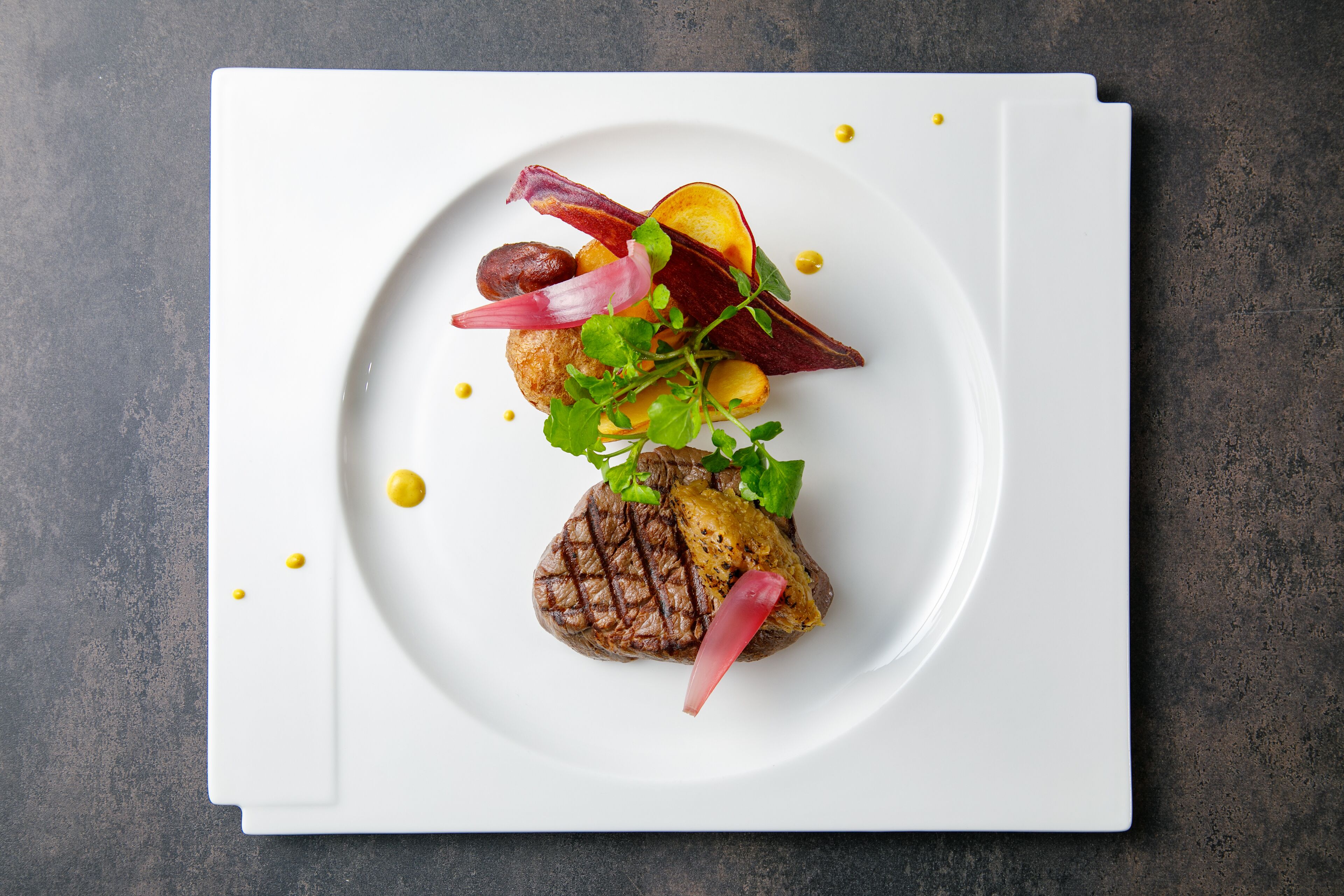 Ein weißer Teller mit gegrilltem Steak, Gemüse und Sauce auf einer dunklen, strukturierten Oberfläche.