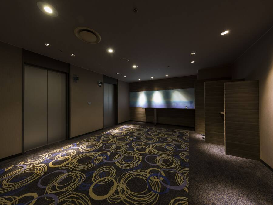 A hotel room with a patterned carpet, a large window with blinds, and a door with a handle.