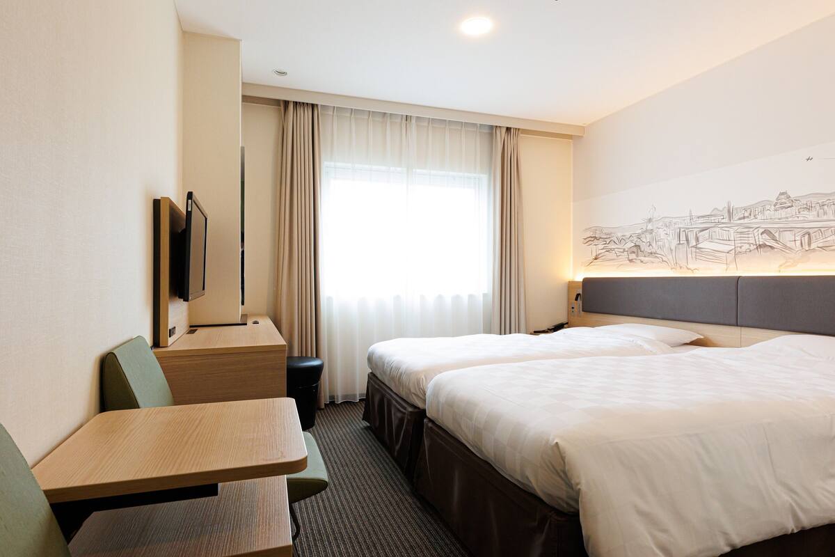 A hotel room with two beds, a desk, a chair, a television, and a window with curtains.