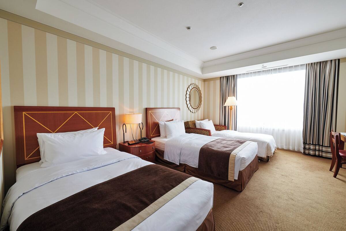 A hotel room with two beds, a large window with curtains, a round mirror on the wall, and a wooden headboard.