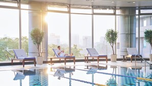 Una piscina interior con agua cristalina, iluminada por amplias ventanas, y sillones de descanso dispuestos alrededor.