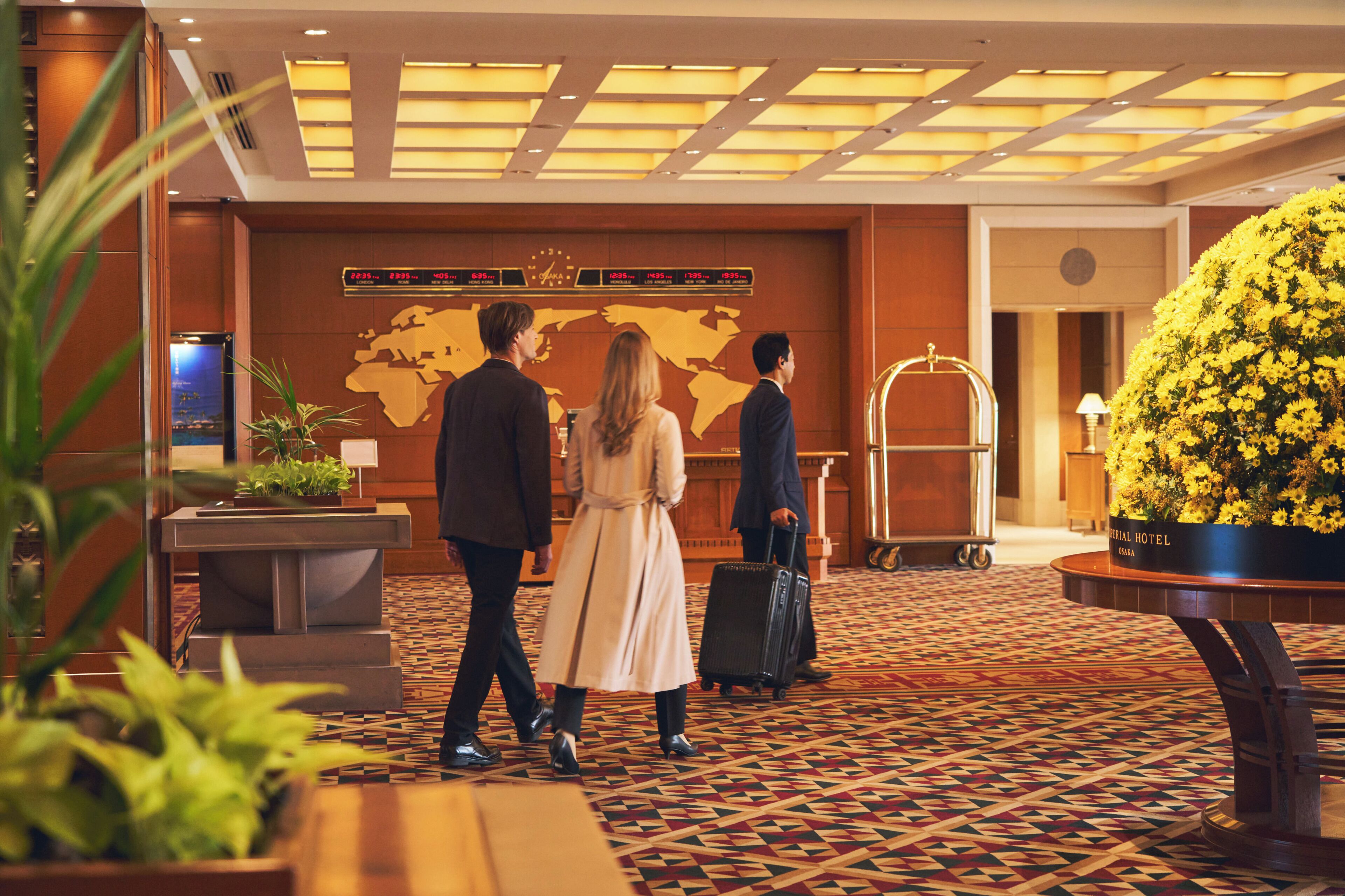 A hotel lobby with a world map, a reception desk, a large flower arrangement, and a luggage cart.