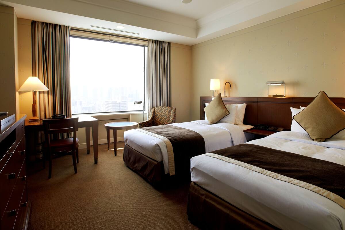 A hotel room with two beds, a desk, a chair, and a window with curtains.