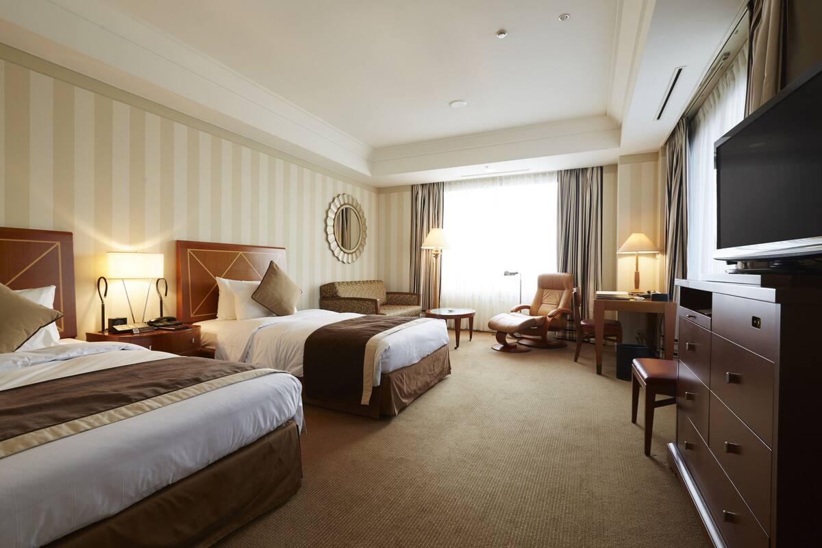 A hotel room with two beds, a desk, a chair, a television, and a dresser.