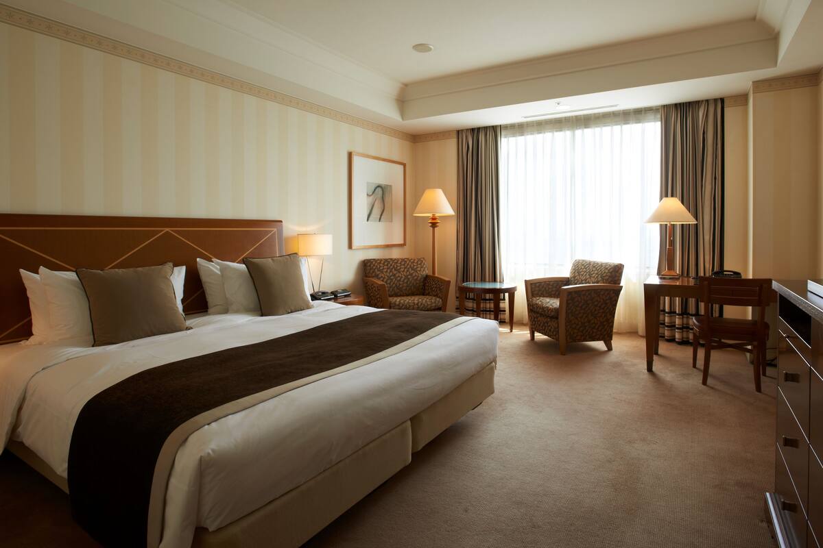 A hotel room with a large bed, two armchairs, a small table, and a desk with a lamp.