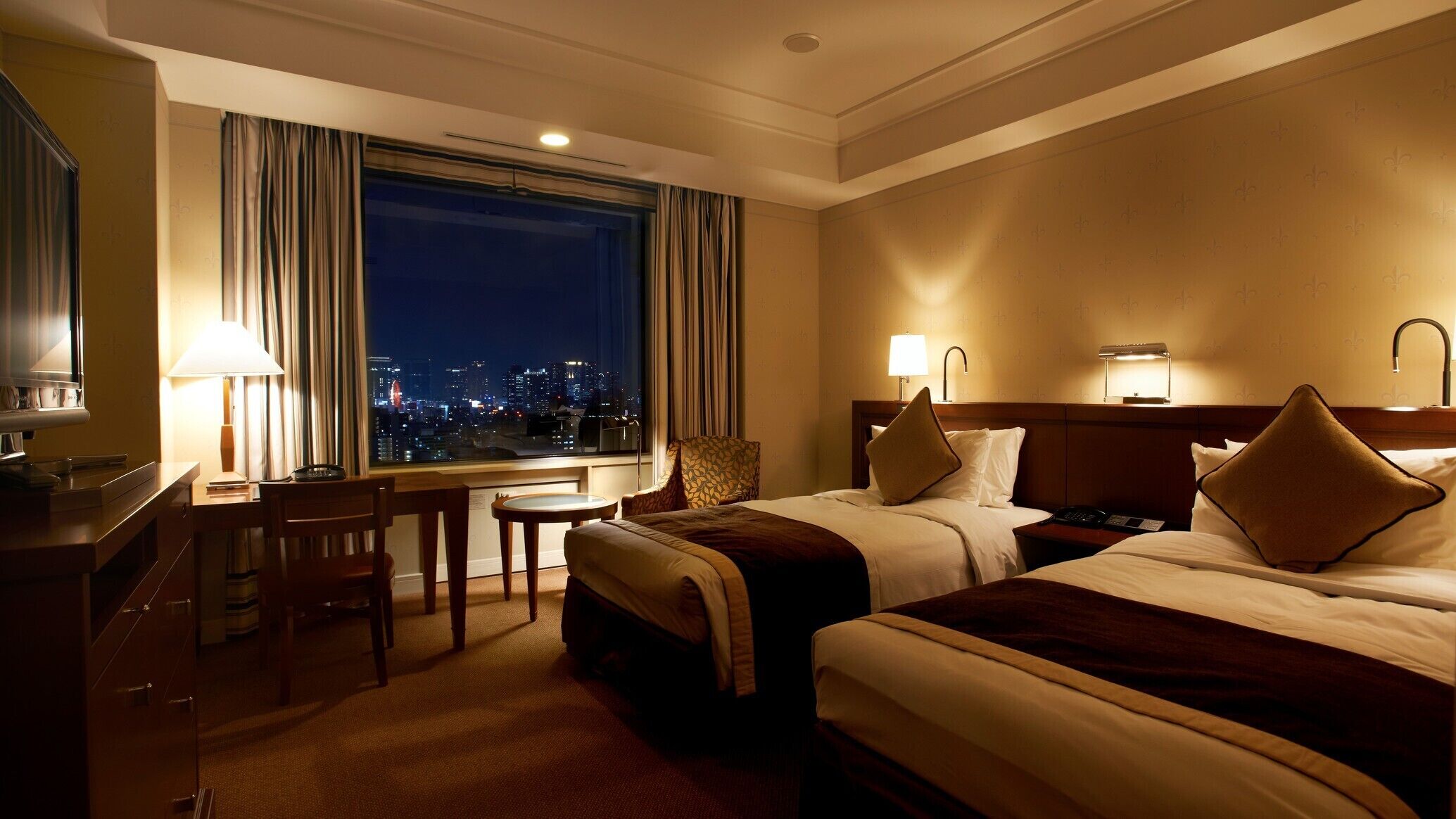 A hotel room with two beds, a desk, a chair, a lamp, and a view of the city at night.